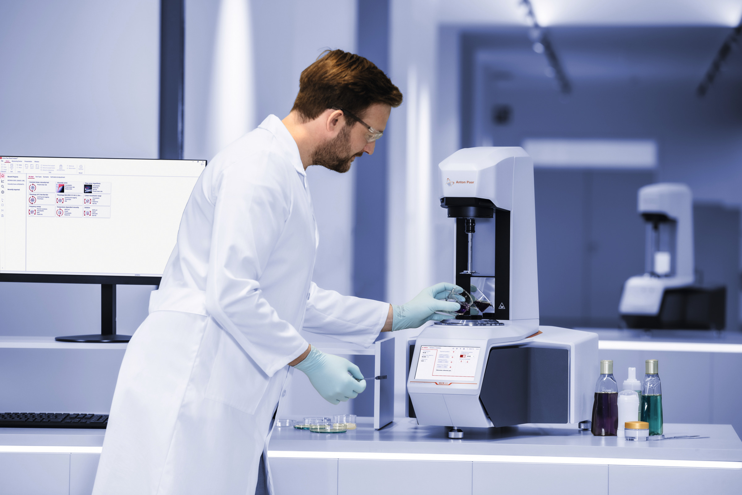 Lab technician in a white coat adjusts a sample on an Anton Paar MCR rheometer beside bottles and a monitor in a laboratory