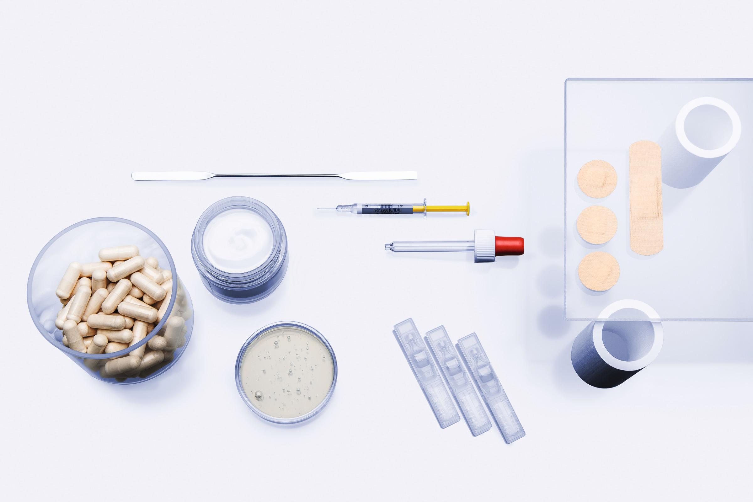 Top-down view of capsules, cream jar, petri dish with gel, syringe, dropper, test strips, and wooden sticks on white surface