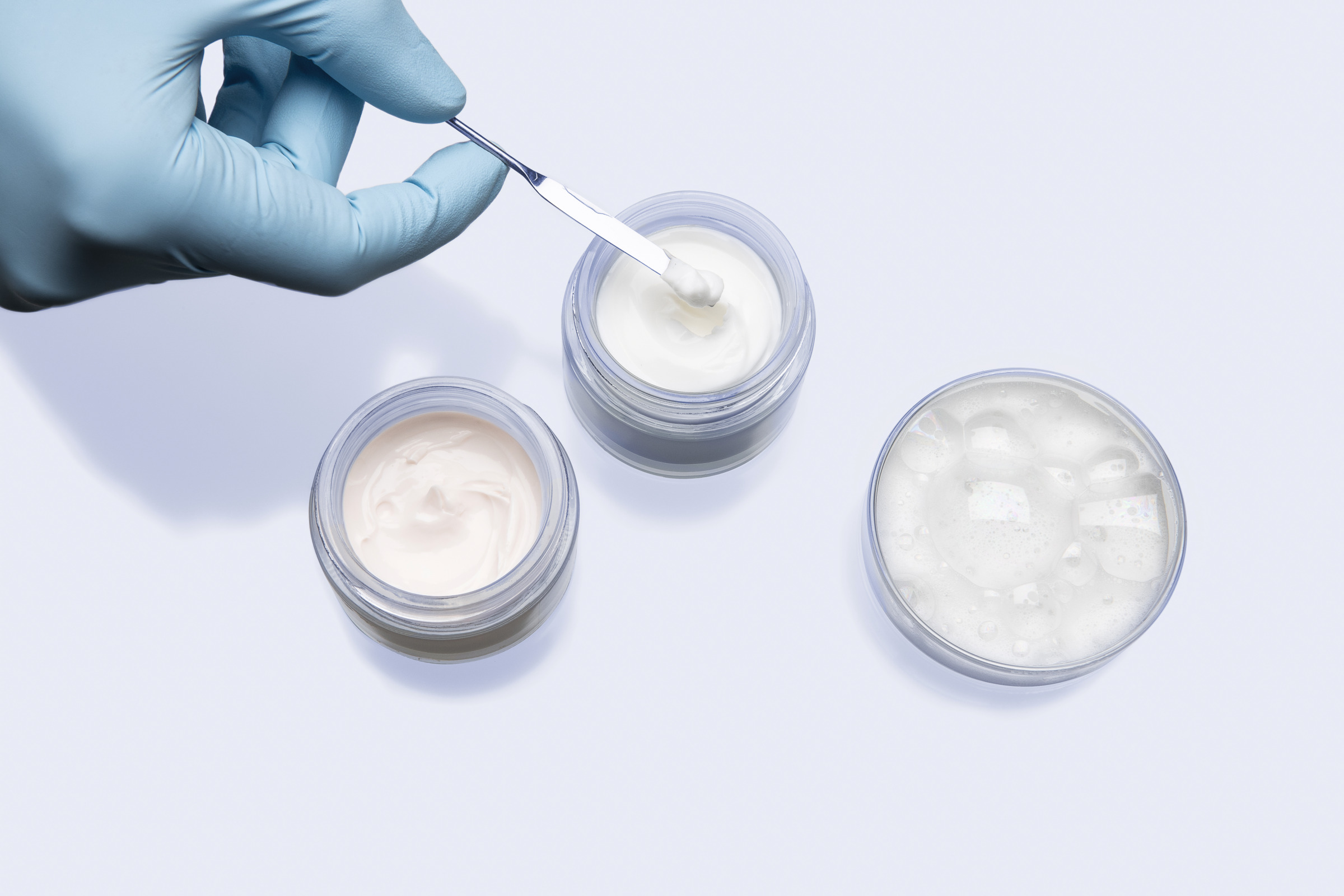 Gloved hand using a spatula to sample cosmetic cream from open jars beside a transparent gel container on a laboratory surface
