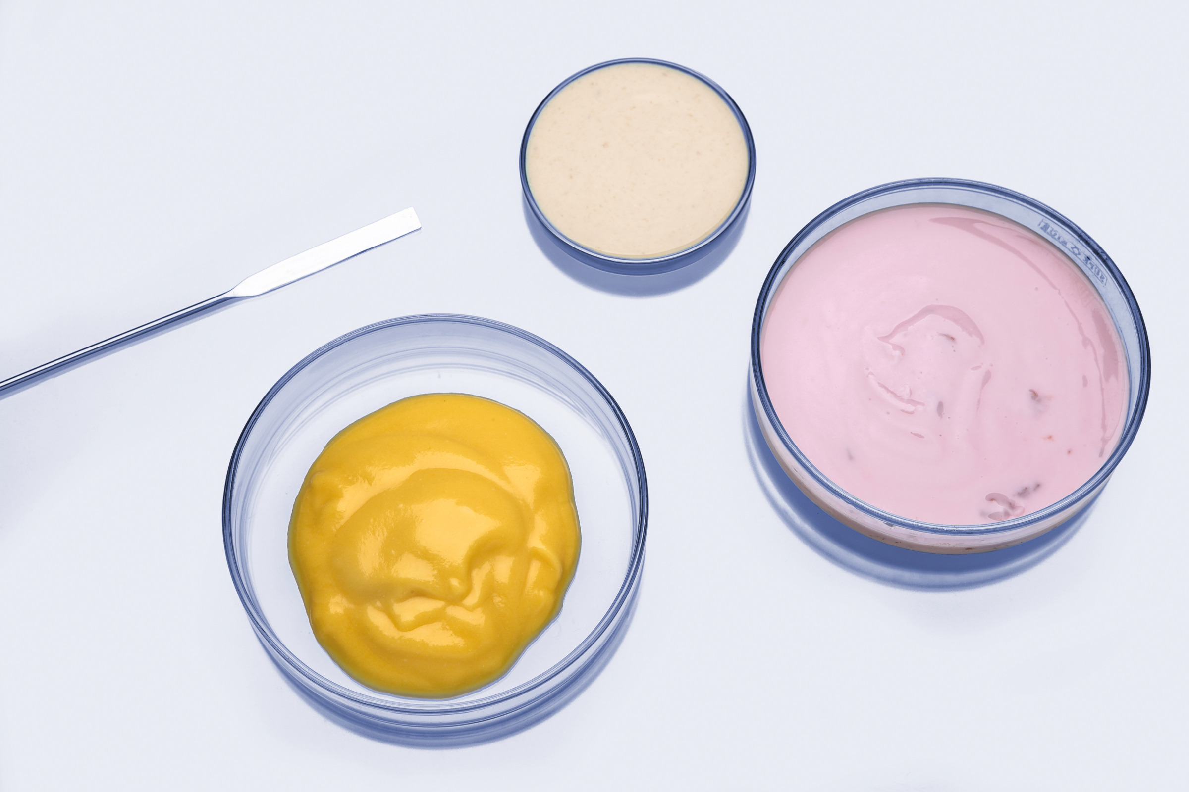 Petri dishes containing yellow, pink, and beige semi-solid food or cosmetic samples with a laboratory spatula on a clean surface