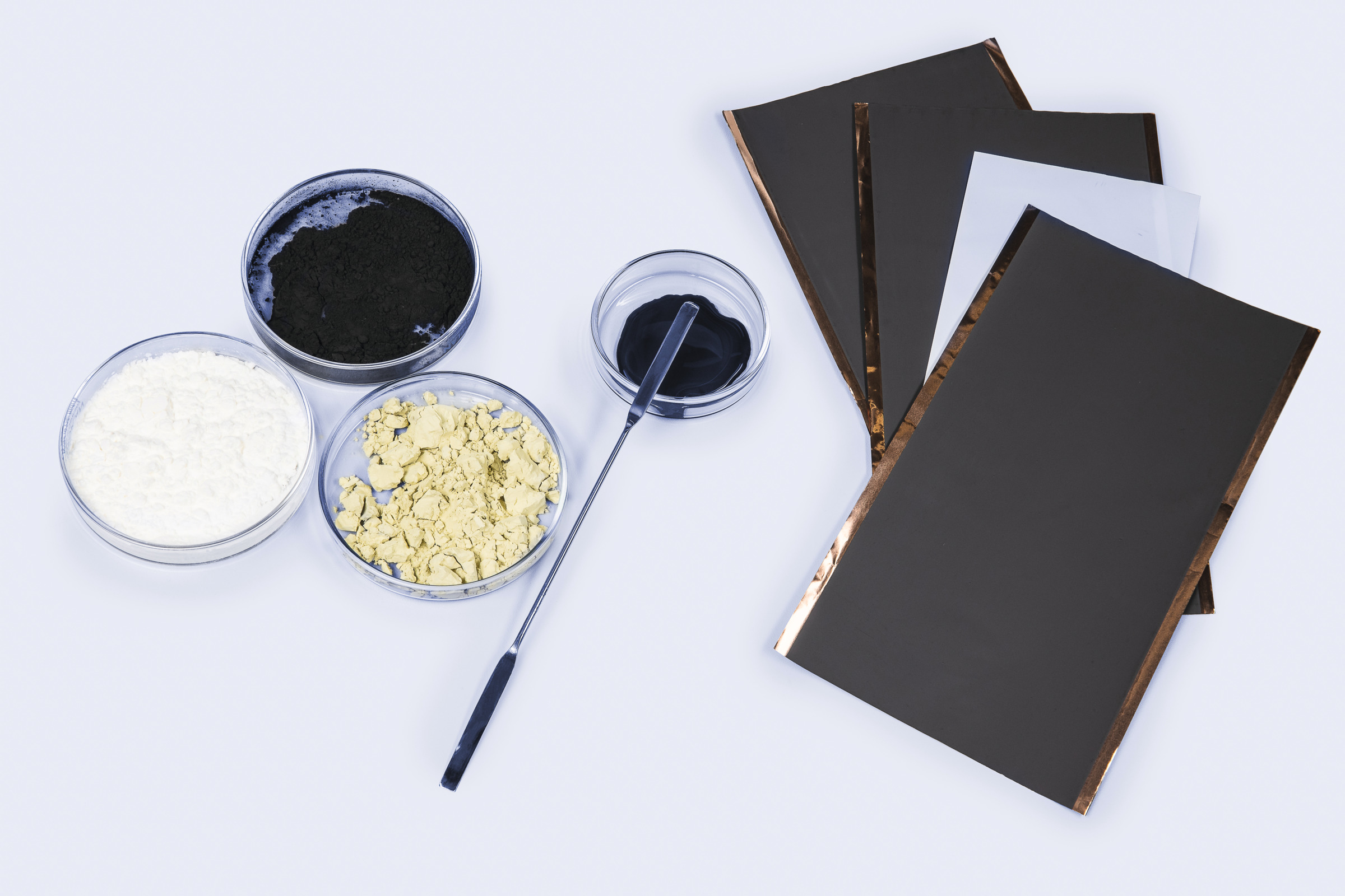 Petri dishes with black, white, and yellow powders, a spatula, and stacked dark electrode sheets with copper-colored edges