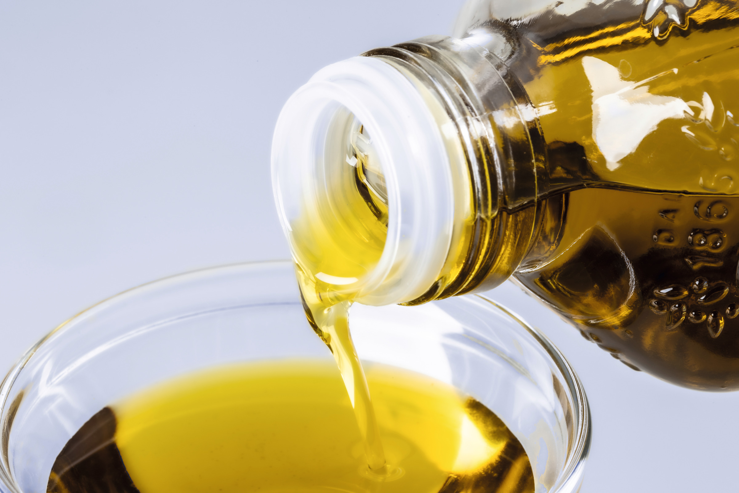 Golden liquid being poured from a glass bottle into a clear dish on a light background