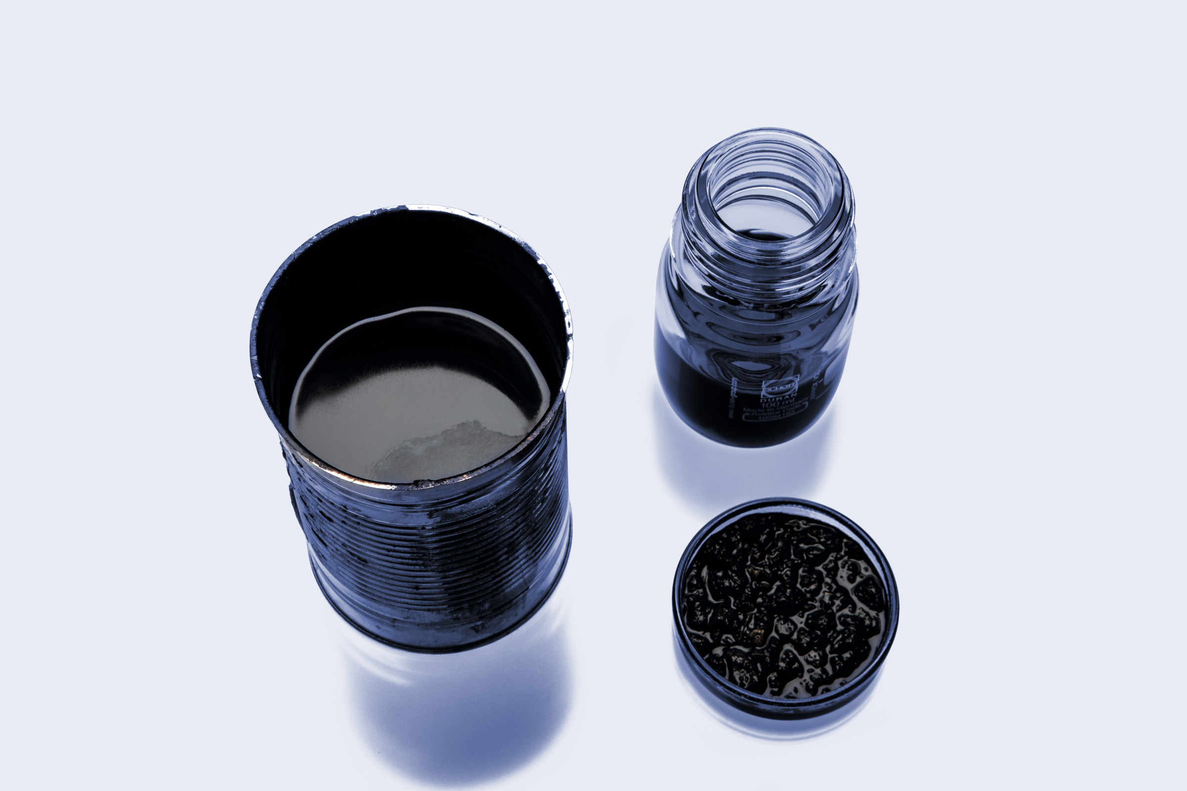 Open metal can, glass bottle, and lid containing black liquid on a light background