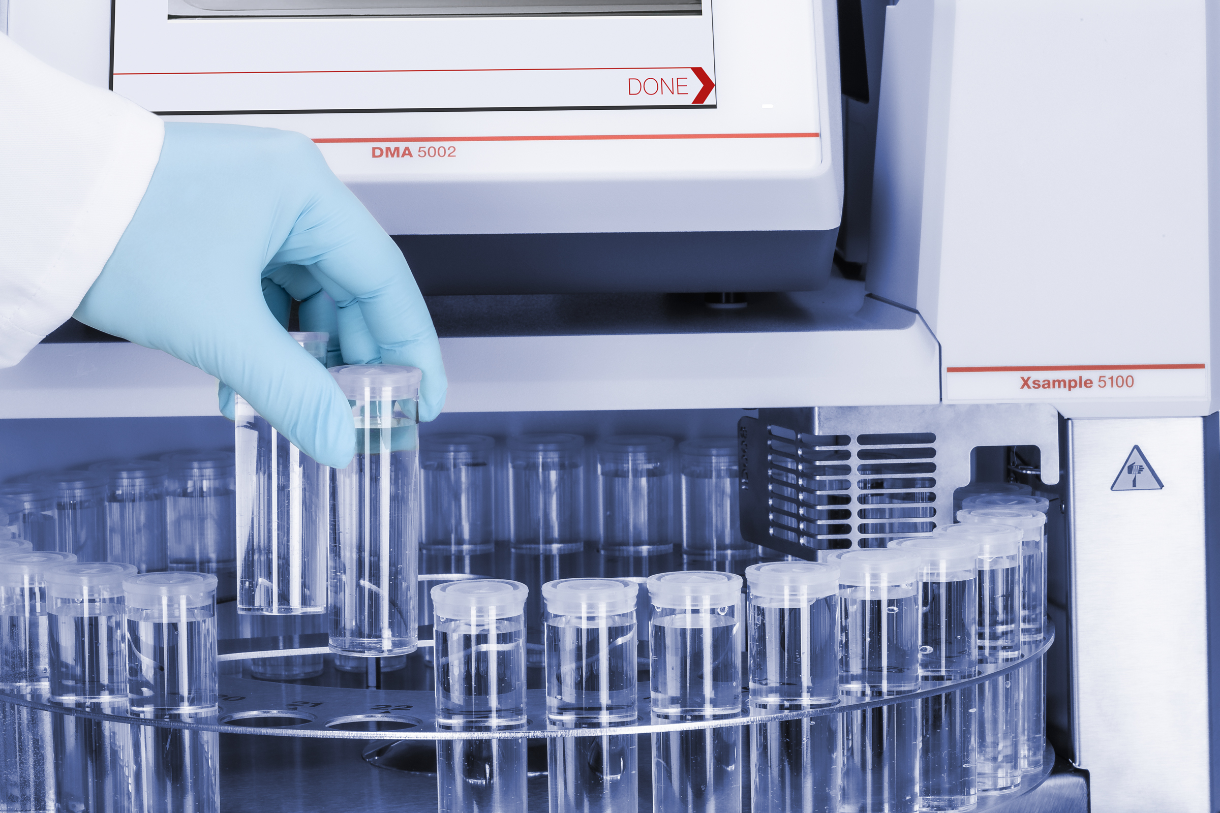 Gloved technician placing a vial in the Anton Paar Xsample beside a DMA 5002 density meter.