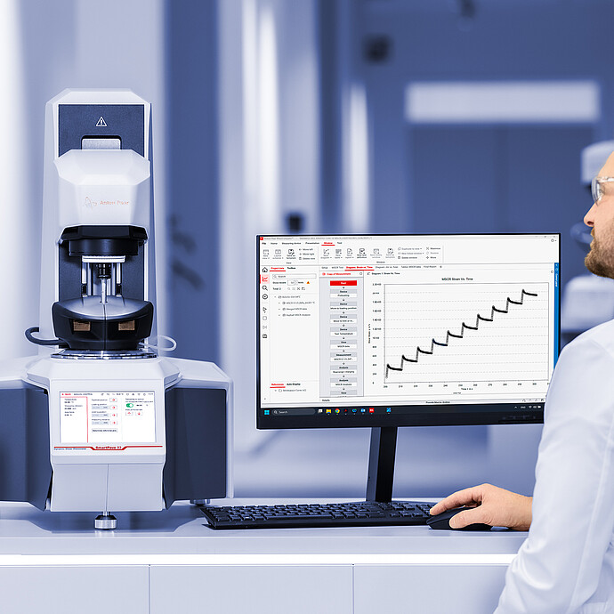 Lab technician operating an Anton Paar SmartPave 93 rheometer, with asphalt samples and a monitor showing RheoCompass strain-time data.