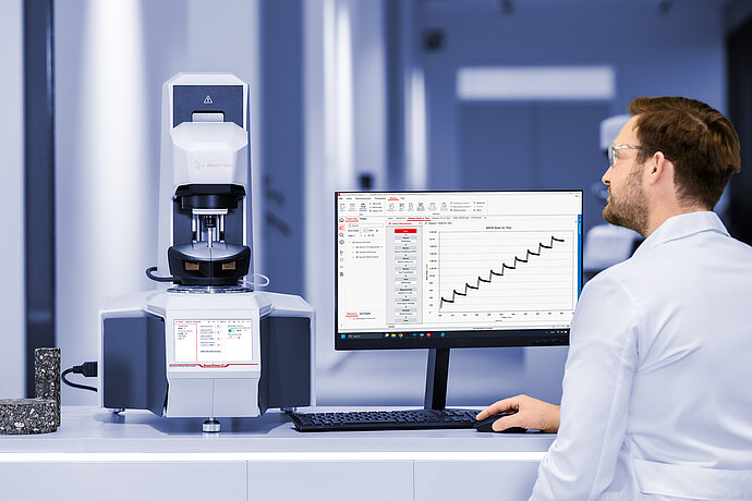 Lab technician operating an Anton Paar SmartPave 93 rheometer, with asphalt samples and a monitor showing RheoCompass strain-time data.