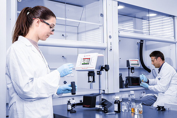Wide lab shot of two technicians working with ViscoQC 100 and ViscoQC 300 rotational viscometers with the PTD 80 Peltier temperature device, with sample bottles and jars on the bench under a fume hood