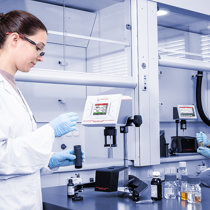 Wide lab shot of two technicians working with ViscoQC 100 and ViscoQC 300 rotational viscometers with the PTD 80 Peltier temperature device, with sample bottles and jars on the bench under a fume hood