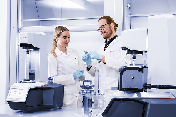 Two lab technicians examining a component between two Anton Paar MCR 503 rheometers in a laboratory setup with measurement accessories.