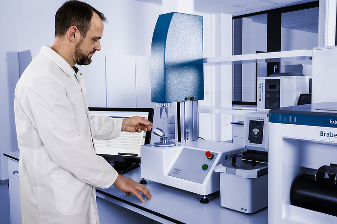 Lab technician operating the Brabender GlutoPeak gluten quality analyzer, loading a sample into the mixing chamber with Julabo unit connected.