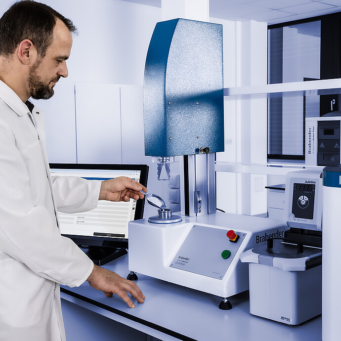 Lab technician operating the Brabender GlutoPeak gluten quality analyzer, loading a sample into the mixing chamber with Julabo unit connected.