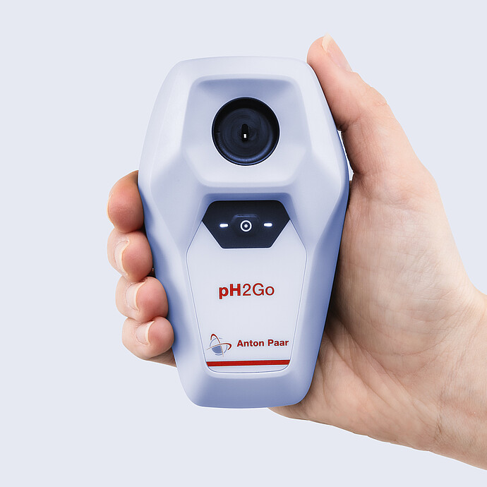 Hand holding an Anton Paar pH2Go digital pH meter, showing the circular sample well, illuminated control button, and compact handheld design.