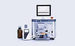 Brabender AbsorptoMeter with hopper and integrated display showing measurement curves, connected via tubing to a dosing unit and reagent bottle on a lab bench