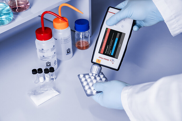 Gloved hands using the Anton Paar Cora 101 handheld Raman spectrometer to analyze tablets in a blister pack on a lab bench with chemicals.