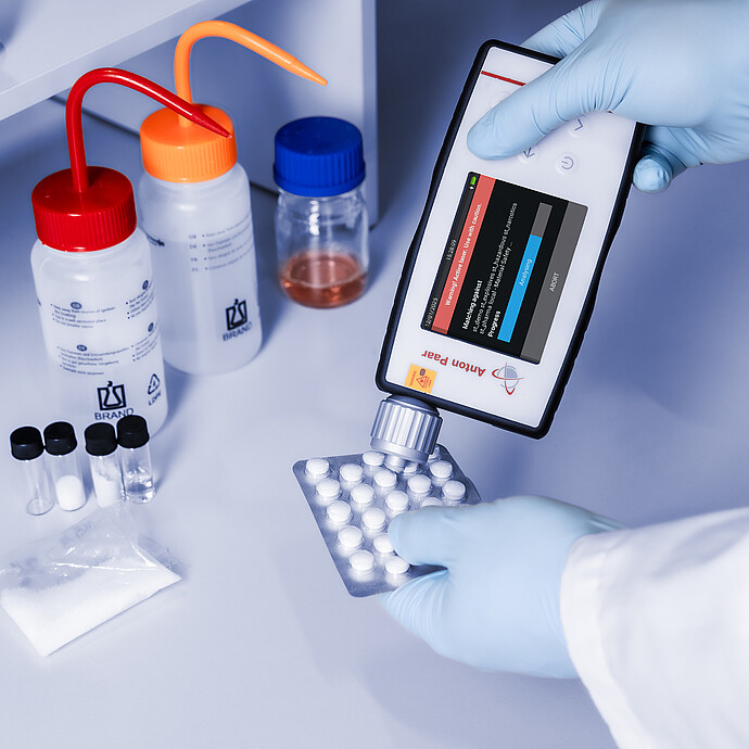 Gloved hands using the Anton Paar Cora 101 handheld Raman spectrometer to analyze tablets in a blister pack on a lab bench with chemicals.