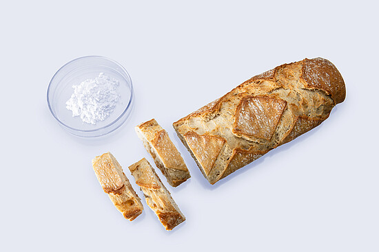 Sliced baguette and white powder in a petri dish arranged on a white surface for bakery sample testing.
