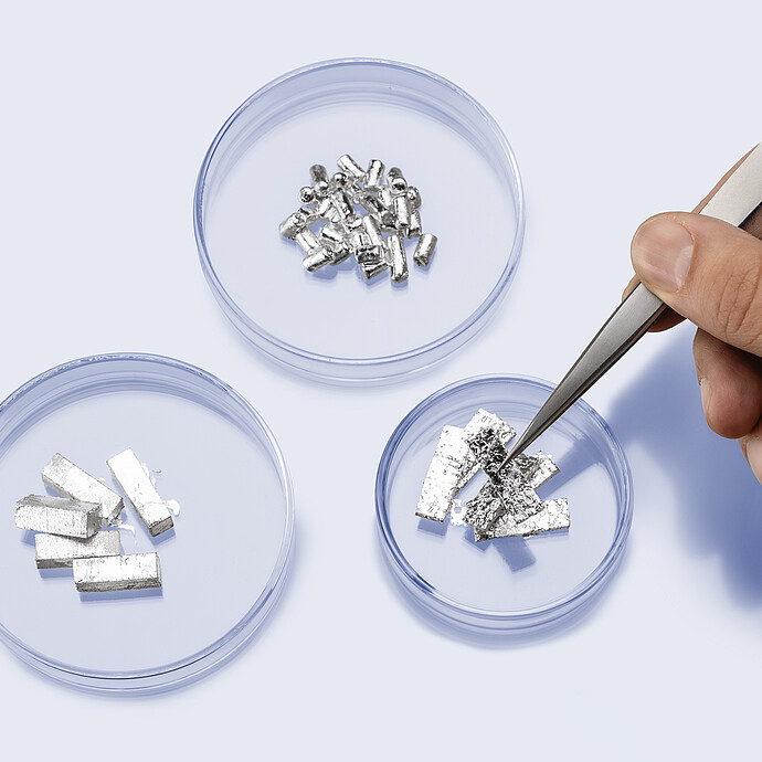 Hand with tweezers handling eGaIn foil in a petri dish, next to additional dishes containing eGaIn pellets and blocks, showing different solid forms of recyclable liquid metal for porosimetry.