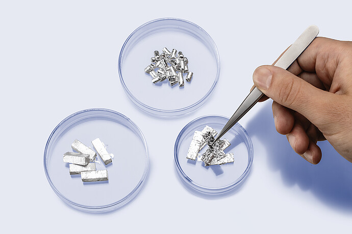 Hand with tweezers handling eGaIn foil in a petri dish, next to additional dishes containing eGaIn pellets and blocks, showing different solid forms of recyclable liquid metal for porosimetry.