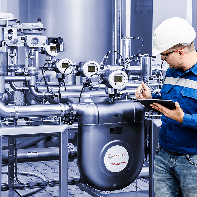 Anton Paar L-Cor 8000 Coriolis flow meter installed in stainless steel piping system, technician in safety gear recording data on a tablet.