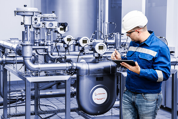 Anton Paar L-Cor 8000 Coriolis flow meter installed in stainless steel piping system, technician in safety gear recording data on a tablet.