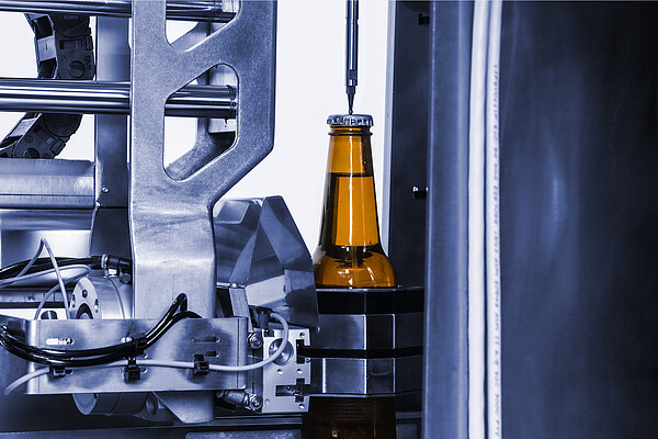 Brown glass beer bottle positioned in an automated production line while a probe measures through the crown cap, surrounded by stainless steel machinery