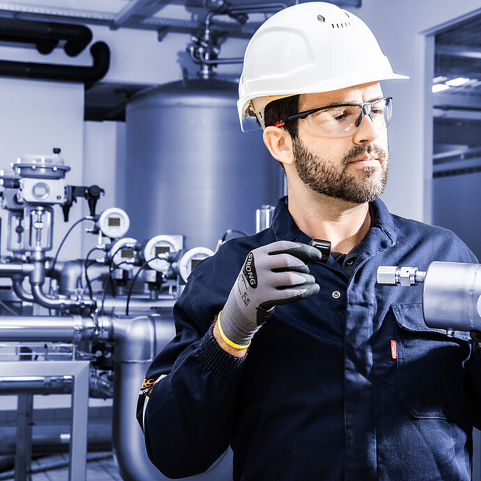 Technician wearing hard hat and safety glasses holding L-Cor 8500 Coriolis flow meter in front of industrial piping and process equipment