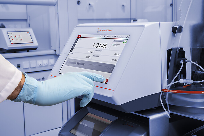 A gloved hand operates the touchscreen of an Anton Paar DMA 4500 M density meter with Xsample 530 sample changer in a lab setup.
