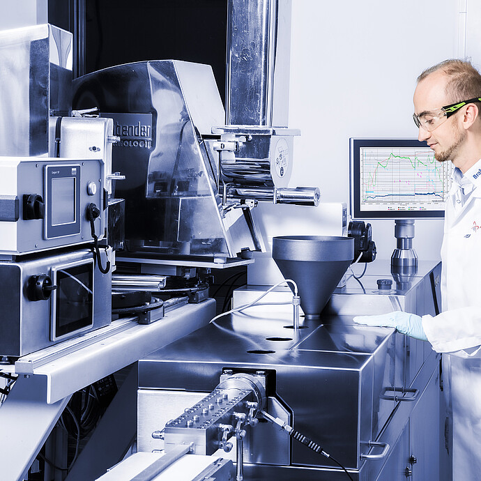 Lab worker in safety glasses beside Brabender TwinLab Series extruder with hopper, die head, monitor and connected feeder unit.