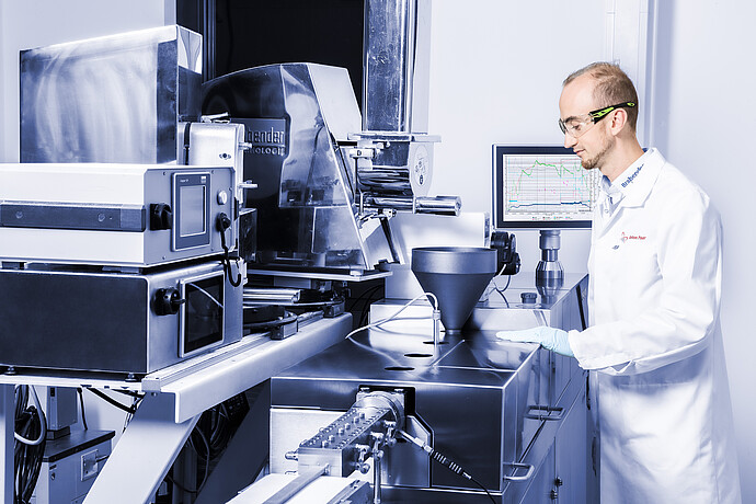 Lab worker in safety glasses beside Brabender TwinLab Series extruder with hopper, die head, monitor and connected feeder unit.