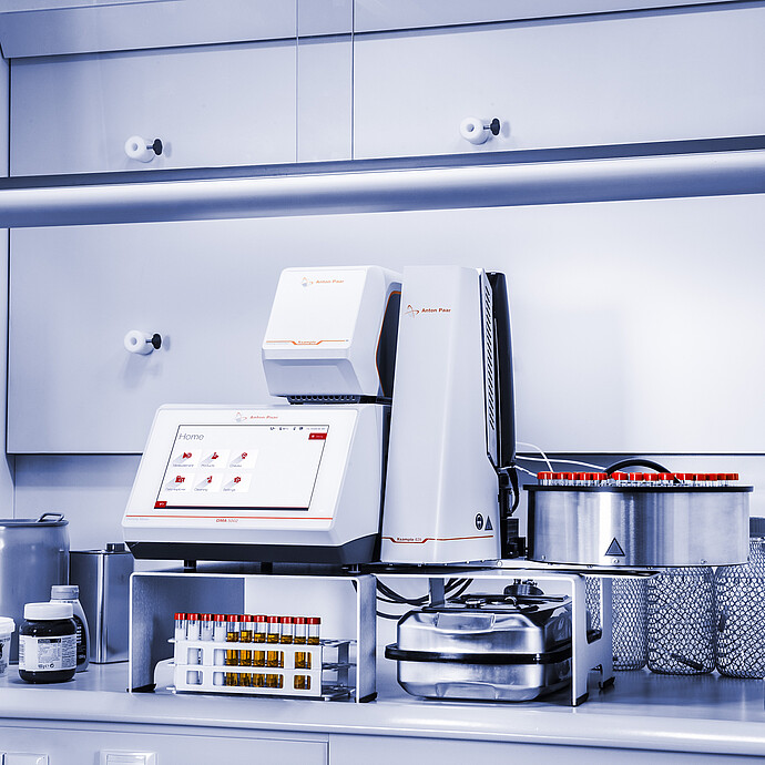 Anton Paar DMA 5002 density meter with Xsample Heating Controller and Xsample 631 sample changer in a fume hood, with red-capped vial carousel, racks, bottles, and containers