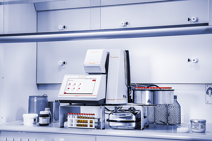 Anton Paar DMA 5002 density meter with Xsample Heating Controller and Xsample 631 sample changer in a fume hood, with red-capped vial carousel, racks, bottles, and containers