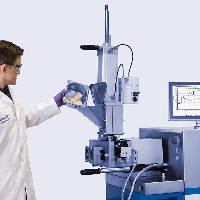 Laboratory technician pours sample pieces into the feed hopper of a Brabender MetaStation 4E modular torque rheometer with graph display.