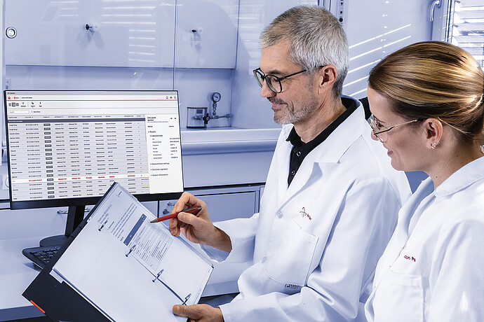 Two Anton Paar lab technicians reviewing measurement data in a laboratory, with AP Connect software open on a monitor and printed result sheets in a folder for comparison and documentation.