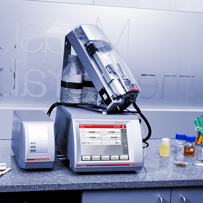 Anton Paar SVM 3001 viscometer with Xsample 610 sample changer and Xsample Heating Controller on a lab bench with bottles, syringes, gloves, and safety glasses