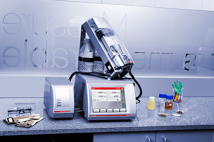 Anton Paar SVM 3001 viscometer with Xsample 610 sample changer and Xsample Heating Controller on a lab bench with bottles, syringes, gloves, and safety glasses