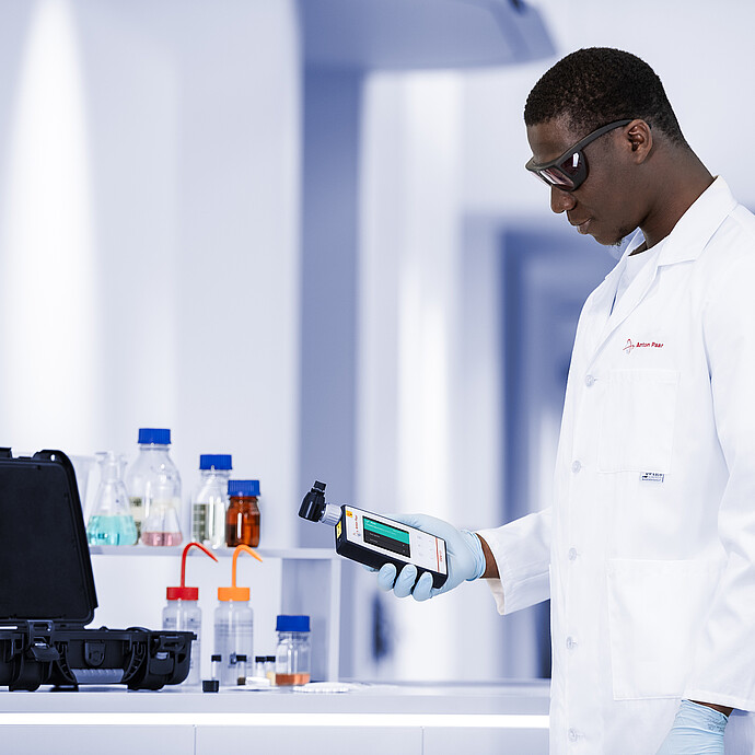 Lab technician using the Anton Paar Cora 101 handheld Raman spectrometer near a workbench with chemicals and an open carrying case.