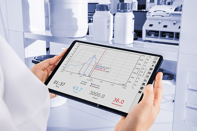 Lab technician holding a tablet displaying Brabender GlutoPeak analysis data with torque, speed, and temperature curves in a lab setting.