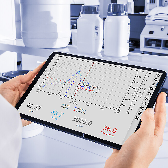 Lab technician holding a tablet displaying Brabender GlutoPeak analysis data with torque, speed, and temperature curves in a lab setting.