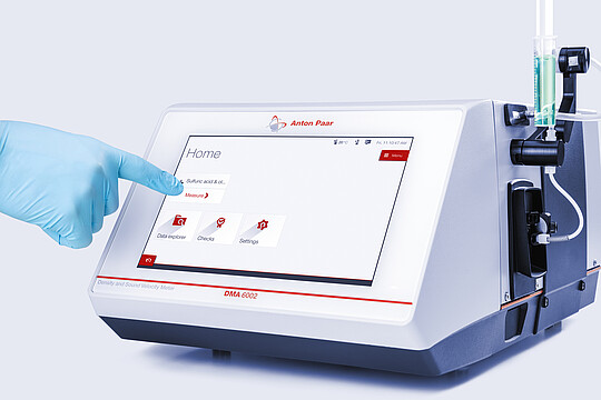 Anton Paar DMA 6002 Density and Sound Velocity Meter with touchscreen, operated by gloved hand; syringe sample inlet attached.