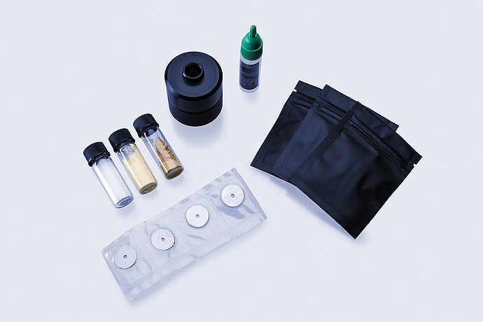 Assorted reference materials including vials with powders, sealed black pouches, a dropper bottle, a sampling accessory, and circular calibration targets.