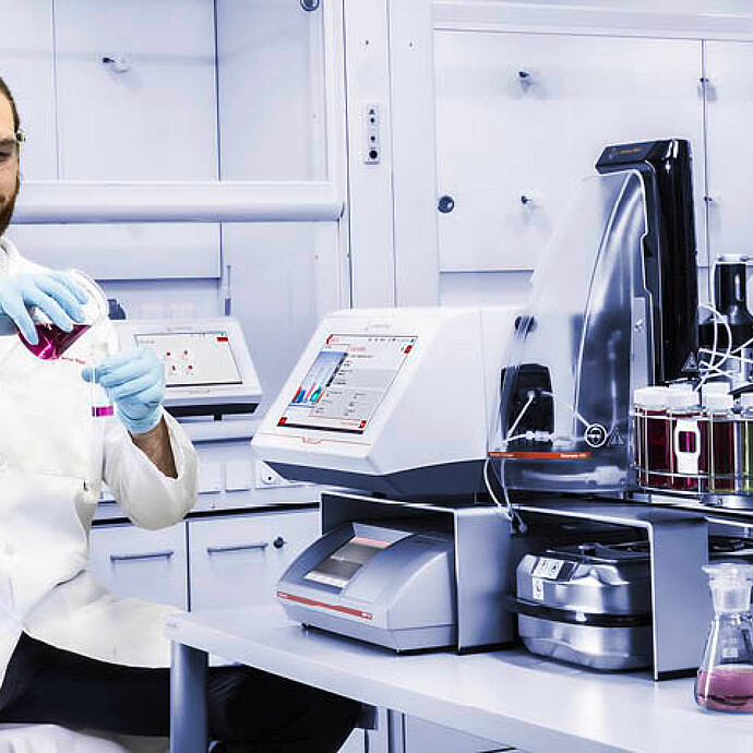 A lab technician prepares a sample next to an Anton Paar DMA 4500 M density meter with Xsample 530 sample changer and label printer.