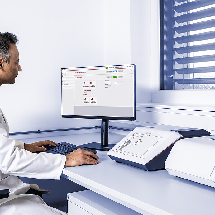 A laboratory technician working at a desk with an Cora 5001 Raman spectrometer on the left and Lyza 3000 FTIR spectrometer on the right, connected to an external monitor displaying the Spectroscopy Suite