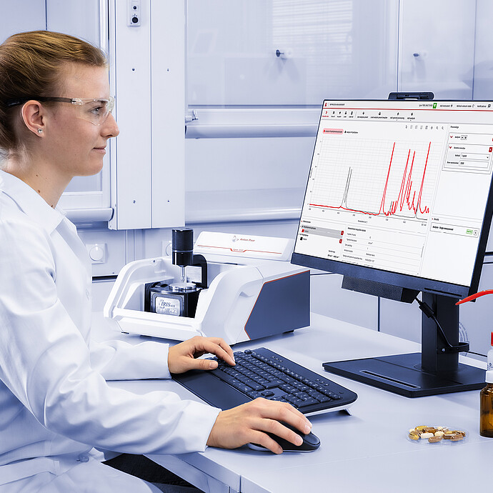 A laboratory technician using an Lyza 3000 FTIR spectrometer to analyze a sample, with an external monitor displaying the Spectroscopy Suite software interface and an analysis spectrum