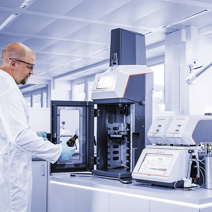 Lab technician loading a beer bottle into the Anton Paar TPO 5001; DMA 4501 M and PFD filling device visible in the background.