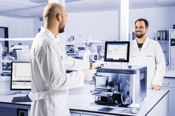 Brabender AbsorptoMeter with open hopper and measuring chamber operated by two lab technicians, integrated display and external monitor visible in laboratory setting