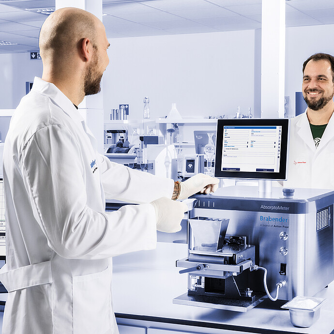 Brabender AbsorptoMeter with open hopper and measuring chamber operated by two lab technicians, integrated display and external monitor visible in laboratory setting