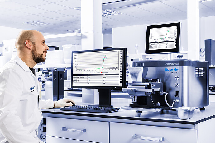 Brabender AbsorptoMeter on lab bench with open hopper and measuring chamber, operated via external monitor displaying torque curve, technician using keyboard