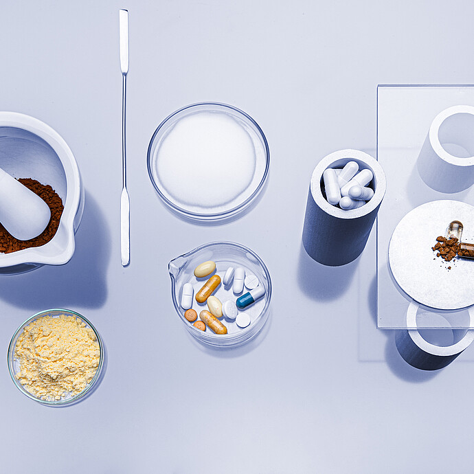 Pharmaceutical materials on a lab bench, including a mortar with powder, a spatula, tablets, capsules, and an opened capsule with contents displayed.