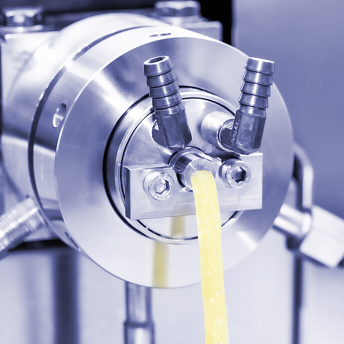 Brabender TwinLab Series extruder die close-up, with yellow plastic strand exiting a circular die head.