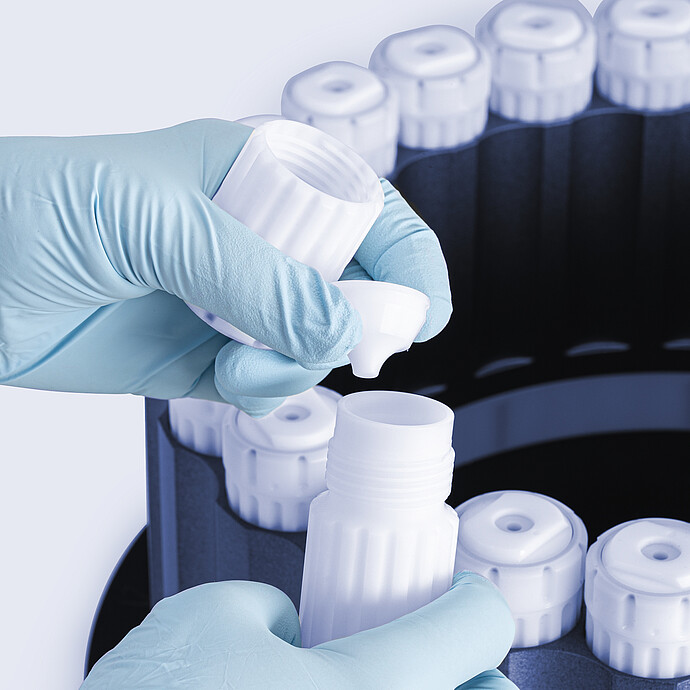 Gloved hands assembling HVT Rotor vessel, placing sealing insert into open digestion vessel, with rotor and capped vessels in the background